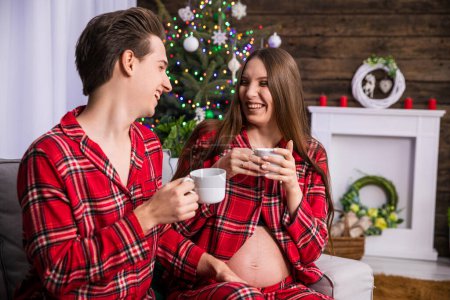 Photo for A man and a woman with advanced pregnancy sit on a couch against the background of a Christmas tree. The couple is holding white teacups in their hands. Both are facing each other, they are cheerful - Royalty Free Image