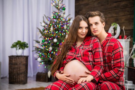 Téléchargez les photos : Une femme enceinte et son mari sont assis ensemble en pyjama rouge. Un sapin de Noël avec des lumières peut être vu en arrière-plan. Deux personnes sourient. - en image libre de droit
