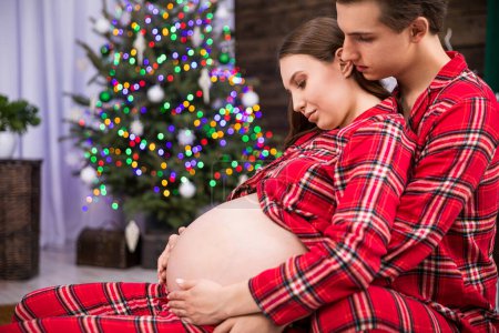 Photo for A pregnant woman leans with her back against a man sitting behind her. The man hugs the woman and holds his hand on her stomach. The hugging couple can be seen in profile against a smudged Christmas - Royalty Free Image