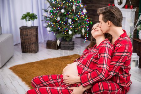 Photo for A pregnant woman has her hands on her stomach. A husband embraces his wife. The man kisses his partner on the cheek. A Christmas tree, a potted plant and a brown carpet can be seen in the background. - Royalty Free Image