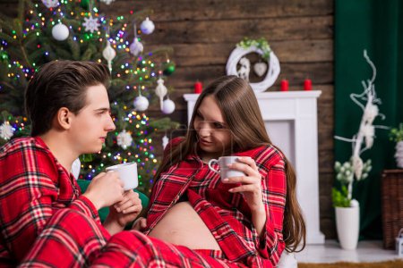 Photo for A couple expecting a baby sits on a gray couch against a backdrop of a dressed Christmas tree with lights. The man is holding the woman on his lap, both are holding white cups and looking into each - Royalty Free Image