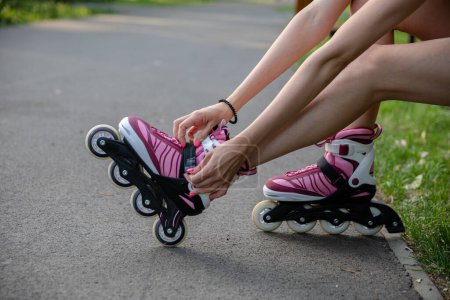 Photo for Close-up of a girls legs putting on rollerblades. Correcting the buckle in rollerblades. The athletic legs of a young person sitting on a park bench. Person in summer short clothing. - Royalty Free Image