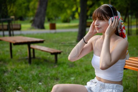 Photo for The girl puts headphones on her head. The woman is styled in a ponytail tied with a colorful scarf. She has pink fingernails. A young person sits in a park, surrounded by trees and greenery. - Royalty Free Image