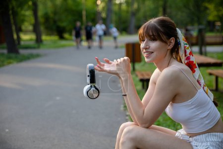 Photo for A happy girl sits on a park bench. She is holding wireless headphones in front of her. There are lots of trees and greenery around. In the distance you can see fuzzy figures of people walking. - Royalty Free Image