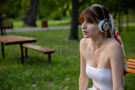 Photo for Woman in the park with wireless headphones on her head. Girl relaxes in the park listening to music. Relaxing in nature in summer. Woman in white strapless top. - Royalty Free Image