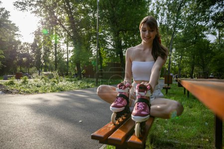 Photo for Spending time in the park on a sunny summer day. A girl is sitting on a brown bench with her legs crossed. She is holding a pair of rollerblades in front of her. Lots of trees and grass in the - Royalty Free Image