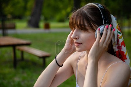 Photo for A close-up of a young girls face. She wears wireless headphones on her head. The woman looks ahead and listens to music. She has a ponytail hairstyle with a long fringe. In the background are fuzzy - Royalty Free Image