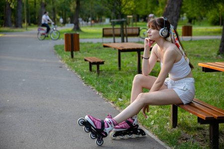 Photo for A thoughtful woman looks ahead while sitting on a bench. She supports her head with her hand. The woman is wearing wireless headphones on her head. In the background you can see a blurry figure riding - Royalty Free Image