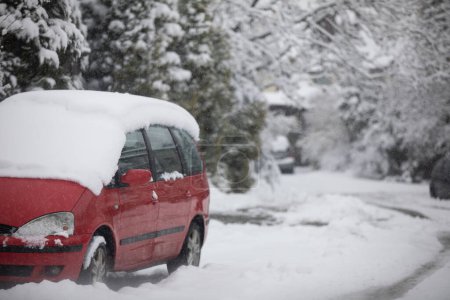 Foto de Un coche rojo de pasajeros es visible en primer plano. El techo del coche está completamente cubierto con una capa de nieve. Fondo borroso. En el fondo se puede ver una calle nevada y copos de caída - Imagen libre de derechos