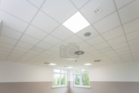 Foto de Techo de panel suspendido visto desde abajo con lámparas encendidas. Justo debajo del techo hay dos ventanas. - Imagen libre de derechos
