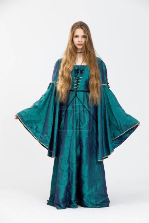 The long-haired girl stands dressed in a long gown to the ground. The dress has flared sleeves. The garment is dark green in color and made of shiny material. The background is white.