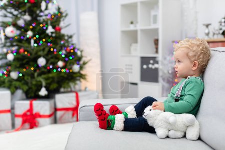 Téléchargez les photos : Un garçon vêtu d'un sweat-shirt vert clair et de bretelles s'assoit sur un canapé gris. À côté de l'enfant se trouve un peluche peluche blanche peluche. En arrière-plan, vous pouvez voir un arbre de Noël décoré de lumières, et - en image libre de droit