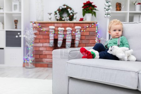 Photo for A young child is in a room decorated with Christmas decorations. In the background you can see a fireplace with lights hanging on it and Christmas gift socks. The boy is sitting on a gray sofa and - Royalty Free Image