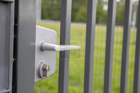 Foto de Una vista de perspectiva de una puerta de acero en una valla con un mango blanco. En el fondo hay un césped verde en un desenfoque suave. - Imagen libre de derechos