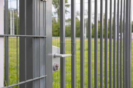 Foto de Una vista de perspectiva de una puerta de acero en una valla con un mango blanco. En el fondo hay un césped verde en un desenfoque suave. - Imagen libre de derechos
