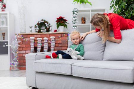 Photo for A woman in a red dress leans over a couch. A little boy is sitting on the couch. The mother is stroking her little sons cheek. Next to the boy lies a plush white toy. - Royalty Free Image