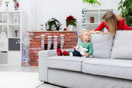 Foto de Un niño pequeño vestido con una sudadera verde claro y tirantes se sienta en un sofá gris. Detrás del sofá hay una mujer vestida de rojo. La mujer se inclina hacia su pequeño hijo. Una chimenea de ladrillo - Imagen libre de derechos