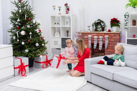 Foto de Una hija y un hijo se alojan con su madre en una habitación decorada con adornos navideños. El niño está sentado en el sofá. La niña está de pie junto al árbol de Navidad, y su madre - Imagen libre de derechos