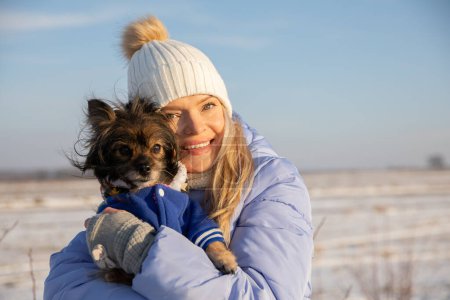 Téléchargez les photos : Une jeune femme souriante serre son petit chien dans ses bras. Le chien est une petite race, de sorte qu'elle peut facilement le porter dans ses bras. La femme et le chien sont dehors un jour d'hiver. - en image libre de droit