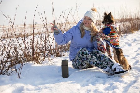 Téléchargez les photos : Une fille est assise dans la neige et pointe du doigt quelque chose. À côté de la fille se tient un chien sur deux pattes. C'est un jour d'hiver lumineux et les deux personnages jettent des ombres dans la neige. - en image libre de droit