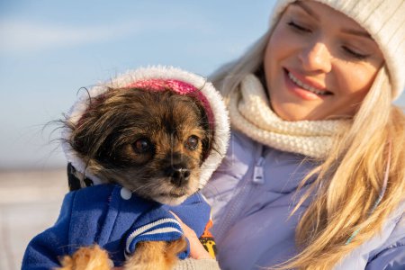 Téléchargez les photos : Une jeune femme blonde sourit en regardant son petit chien. Elle tient le chien dans ses bras. La femme et le chien sont dehors pendant qu'il fait froid. - en image libre de droit