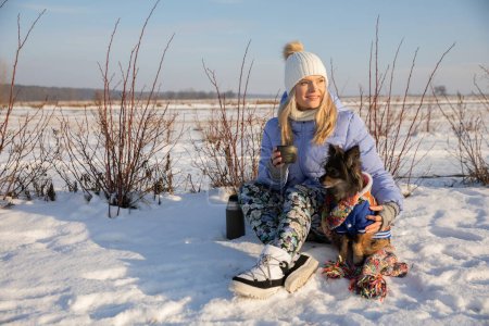Téléchargez les photos : Promenade avec votre chien au coucher du soleil. Un voyage avec un chien hors de la ville dans le désert. Une fille boit du thé chaud dehors en hiver enneigé. - en image libre de droit
