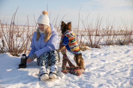Téléchargez les photos : Une fille est assise dans un champ avec son chien. Il neige partout. La fille porte des vêtements chauds : veste, chapeau et bottes de neige. Le chien se tient debout sur deux pattes et s'appuie contre son propriétaire. - en image libre de droit