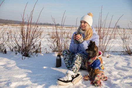 Téléchargez les photos : Une femme se perd en dehors de la ville. Elle s'assoit dans la neige et tente de se réchauffer en buvant une boisson chaude dans un thermos. À côté de son propriétaire se trouve un chien de petite race. - en image libre de droit