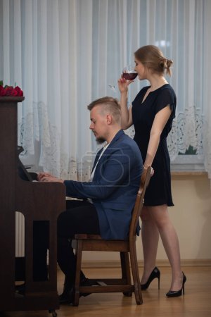 Foto de Un pianista interpreta una pieza de piano de memoria. Al lado del hombre se mueve una mujer joven elegantemente vestido. Bebe vino tinto de una copa.. - Imagen libre de derechos