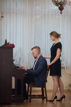 Foto de Una mujer vestida con un elegante vestido negro observa a su marido. El marido toca el piano. El hombre lleva un traje oscuro y una camisa blanca. Hay rosas tiradas en el piano. - Imagen libre de derechos