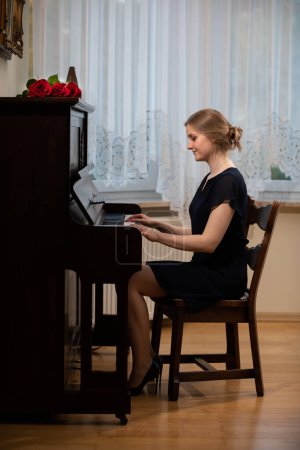Foto de Una mujer elegante toca el piano en la sala de estar. Lleva un vestido negro y tacones altos. Un ramo de rosas rojas se encuentra en el piano. - Imagen libre de derechos