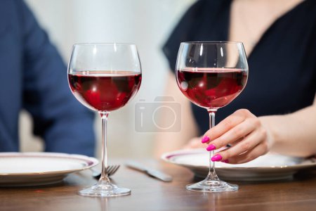 Téléchargez les photos : Deux verres brillants de vin rouge se tiennent sur la table. Près d'eux se trouvent deux petites assiettes. En arrière-plan, vous pouvez voir deux figures floues assis à la table. La femme tient une des lunettes.. - en image libre de droit