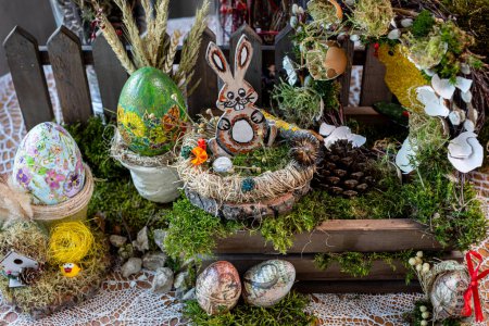 La escena captura una encantadora composición de Pascua vista desde arriba, con una figura de conejo de madera enclavada en un nido de aves sobre una caja de madera. En el fondo, se muestran coloridos huevos de Pascua