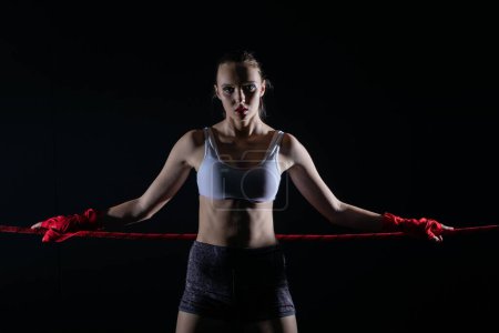 Téléchargez les photos : L'athlète est basé sur la corde rouge du ring. Une femme déterminée se tient sur le ring prête à commencer le combat. - en image libre de droit