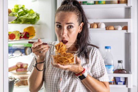 Foto de Asustada mujer hambrienta en pijama está comiendo espaguetis en el refrigerador por la noche. - Imagen libre de derechos