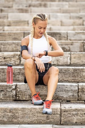 Téléchargez les photos : Femme Runner vérifier les résultats sur les montres intelligentes, assis dans les escaliers, prendre pouls après le jogging et le repos, brassard avec téléphone portable. - en image libre de droit