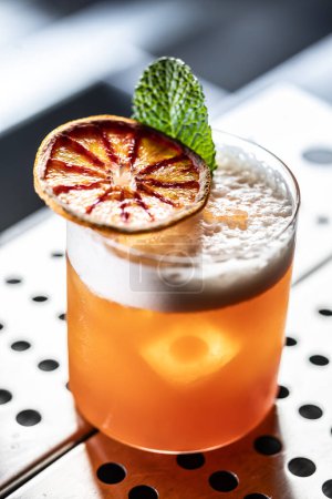 Cóctel de vaso de Jungle Bird adornado con naranja roja seca y menta en el mostrador del bar.