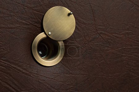 Peephole with an open damper on a brown leatherette door.