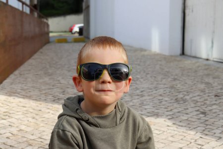 Foto de El rubio de pelo corto sacando los labios y haciendo el tonto. Un niño de cinco años con gafas de sol. - Imagen libre de derechos