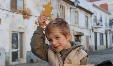 Téléchargez les photos : Un garçon blond tient un cookie en forme de cerf au-dessus de sa tête. Un garçon de cinq ans fait l'idiot et s'amuse.. - en image libre de droit