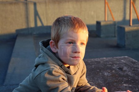 Foto de Un chico rubio mira pensativamente a la distancia mientras está sentado en el techo de una casa al atardecer. - Imagen libre de derechos