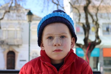 Portrait of a pensive boy in a red jacket and blue hat. A detached six-year-old boy looks into the distance.