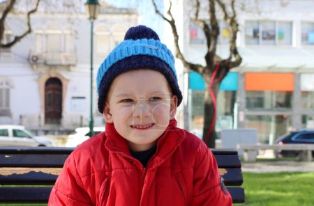 Foto de Un niño con chaqueta roja y sombrero azul se arruga la nariz y se desnuda los dientes. Un niño de seis años hace muecas, mostrando una cara enojada. - Imagen libre de derechos