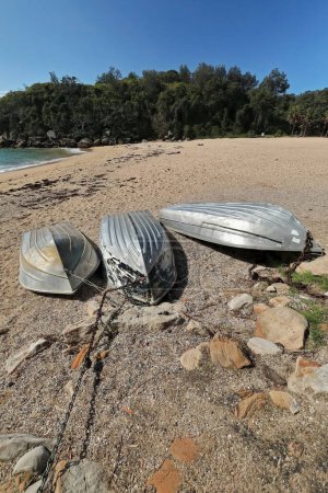 Photo for Upside down beached aluminum boats on the Shelly Beach golden sand and gravel-moored to padlocked rusty chains and cables-grounded next to the Marine Parade walkway. Manly suburb-Sydney-NSW-Australia. - Royalty Free Image