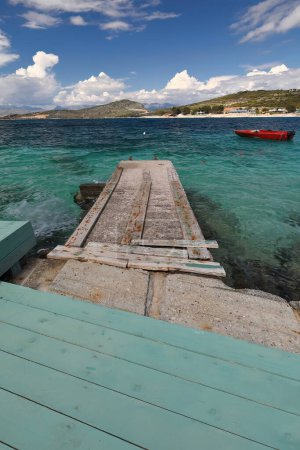 Photo for Cement pier jutting into the sea from a wood green platform on the north-facing Rilinda beach overlooking green-blue turquoise waters, red outboard tourist boat, Ksamil village. Saranda city-Albania. - Royalty Free Image
