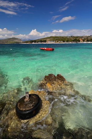 Photo for Mooring made of an old tire cemented to a rock-embedded wooden post, north-facing Rilinda beach overlooking green-blue turquoise water, red outboard tourist boat, Ksamil village. Saranda city-Albania. - Royalty Free Image