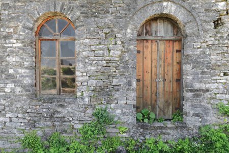 Photo for Chipped, paint-faded, unpainted, padlocked, round arch wood door and glazed window, both cut into the stone wall of an Ottoman-era building in the Clock Tower area of the citadel. Gjirokaster-Albania. - Royalty Free Image