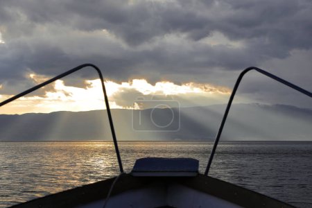 Téléchargez les photos : Rayons lumineux crépusculaires - appelés rayons de Dieu- projetant sur le bord du lac d'Ohrid les montagnes Shebenik-Jabllanica, frontière AL-NMK, vus depuis un bateau naviguant sur le lac en fin d'après-midi pluvieux. Ohrid-Macédoine du Nord. - en image libre de droit