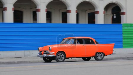 Foto de La Habana, Cuba-07 de octubre de 2019: El viejo auto clásico americano rojo-naranja almendro, tanque de yanqui Dodge Kingsway Custom Sedán de 4 puertas de 1956 conduce a lo largo del paseo marítimo del Prado pasando una valla de chapa metálica. - Imagen libre de derechos