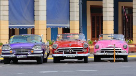 Foto de La Habana, Cuba-07 de octubre de 2019: Coches descapotables clásicos americanos, azul púrpura Dodge Custom Royal Lancer 57-red Ford Crestline Sunliner 52-pink Chevrolet Styleline DeLuxe 52 park on Paseo del Prado. - Imagen libre de derechos